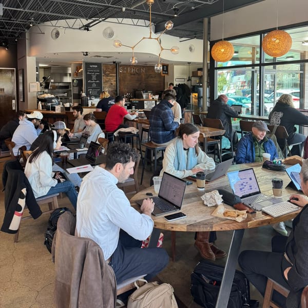 Customers work on laptops at wooden tables in a busy coffee shop with large windows.