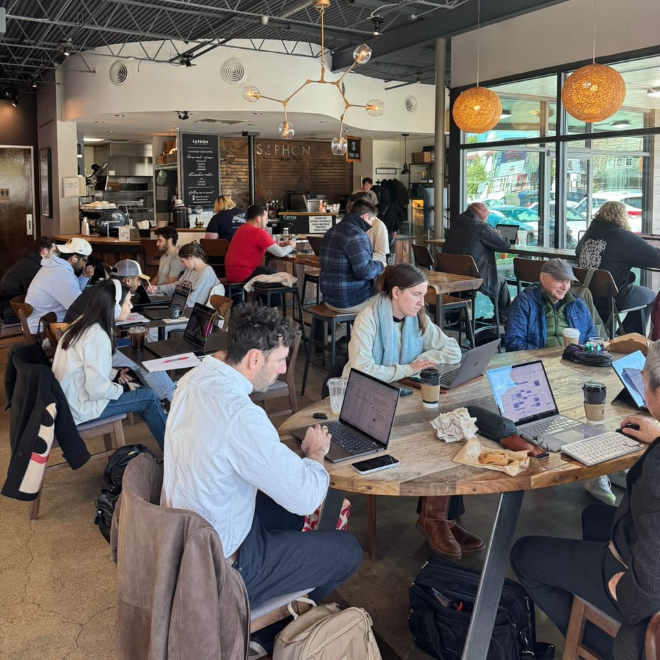 Customers work on laptops at wooden tables in a busy coffee shop with large windows.