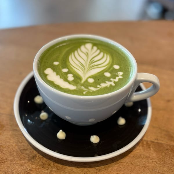 A vibrant green matcha latte with intricate white latte art sits on a black saucer with white dots.