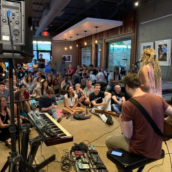A musician plays guitar to an audience seated on the floor, while a singer performs on stage.