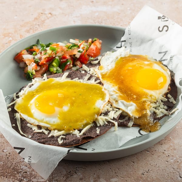 Huevos rancheros with green and yellow sauce, shredded cheese, and pico de gallo on a gray plate.