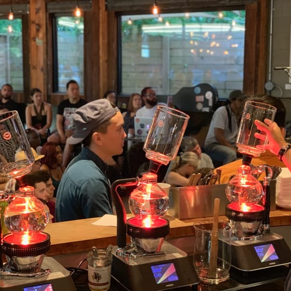 People watching a coffee brewing demonstration with three siphon brewers on a wooden counter.
