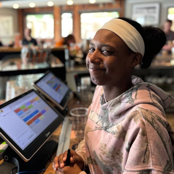Smiling restaurant staff member in a pink camouflage hoodie stands by two point-of-sale screens.