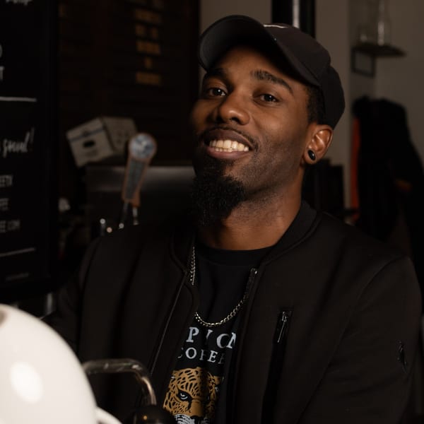 A smiling man in a black cap and jacket with a leopard print shirt looks directly at the camera.