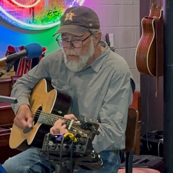 A man with a beard and glasses, wearing a baseball cap, plays an acoustic guitar.