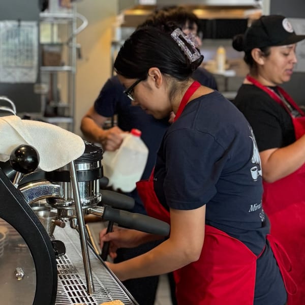 Barista in a red apron and glasses operating an espresso machine, with other staff in the background.