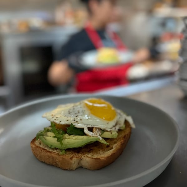 Avocado toast with a fried egg and cilantro on a gray plate, with a chef in the background.