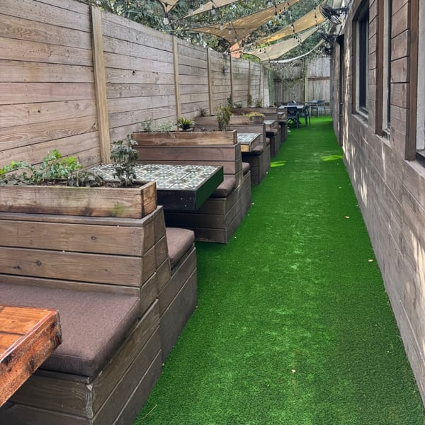 Outdoor restaurant patio with wooden booth seating, green turf, and string lights under shade sails.