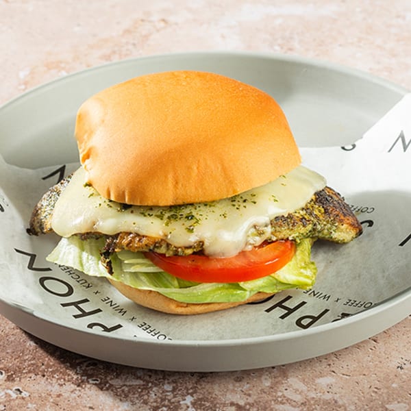 A chicken burger with melted cheese, pesto, lettuce, and tomato on a bun, served on a plate.