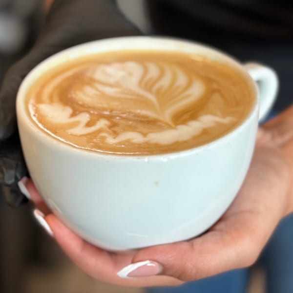 Hands in black gloves holding a white mug with latte art on a dark background.