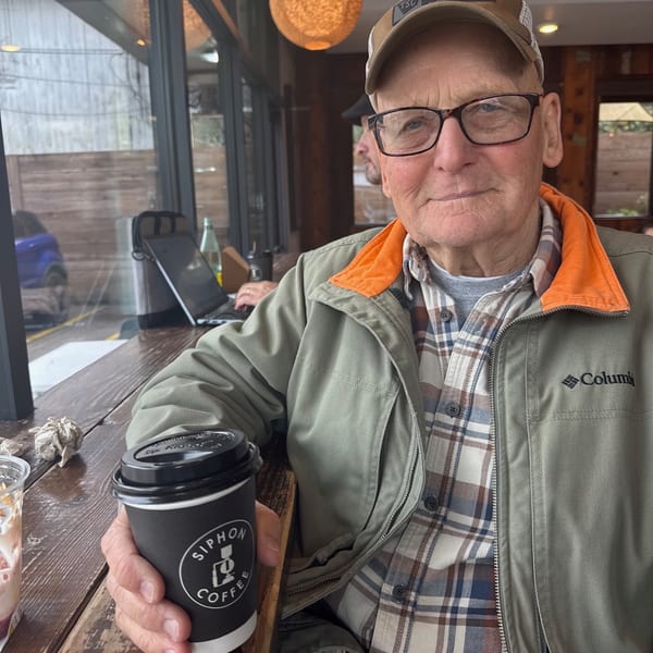 An older man in a cap and glasses holds a Siphon Coffee cup at a wooden table.