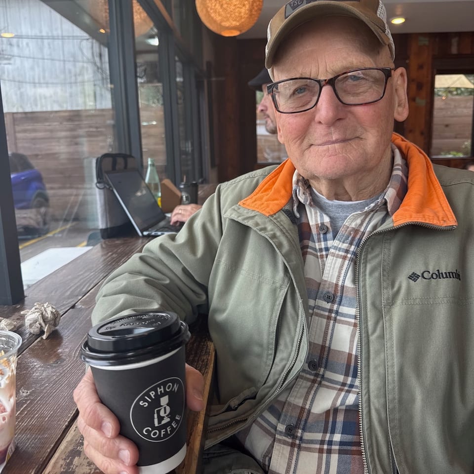 An older man in a cap and glasses holds a Siphon Coffee cup at a wooden table.