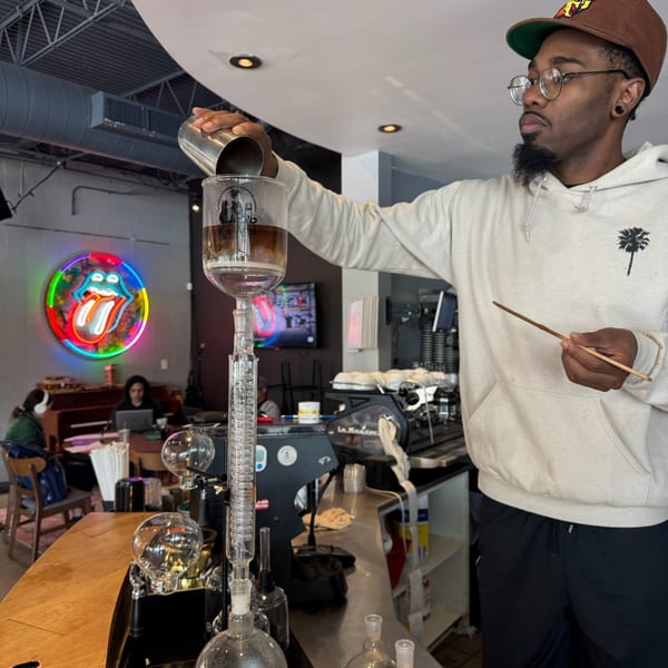 A barista pours liquid into a cold brew coffee maker, with a neon Rolling Stones sign in the background.