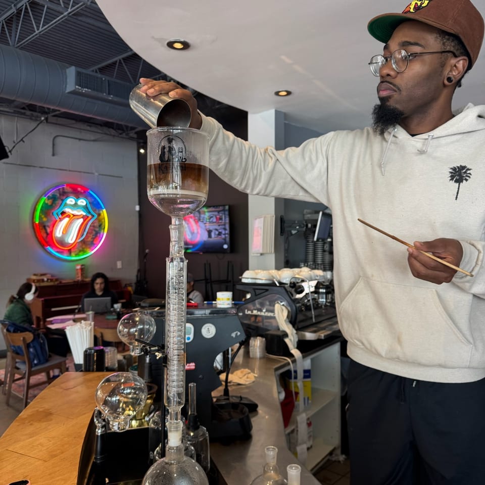 A barista pours liquid into a cold brew coffee maker, with a neon Rolling Stones sign in the background.