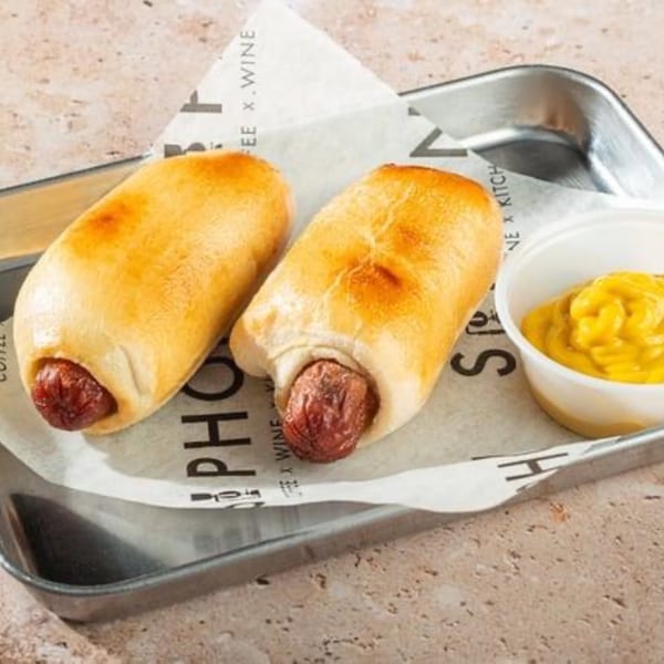 Two golden-brown pigs in a blanket with a side of yellow mustard on a metal tray.