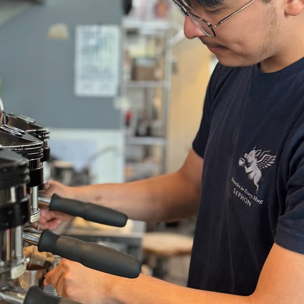 Barista in glasses and a dark t-shirt with a winged pig logo operating an espresso machine.