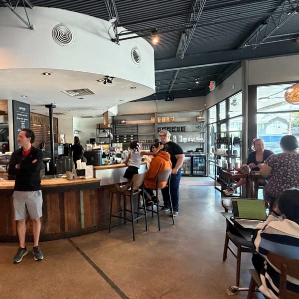 Customers and staff inside a modern coffee shop with a wooden counter and large windows.