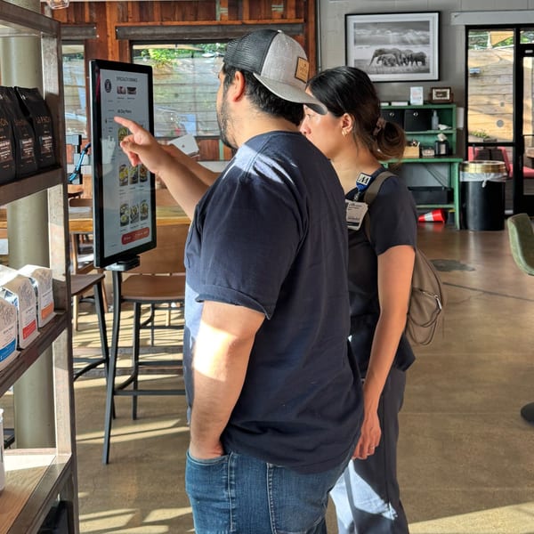 Two people ordering from a digital menu kiosk in a sunlit cafe with coffee bags on shelves.