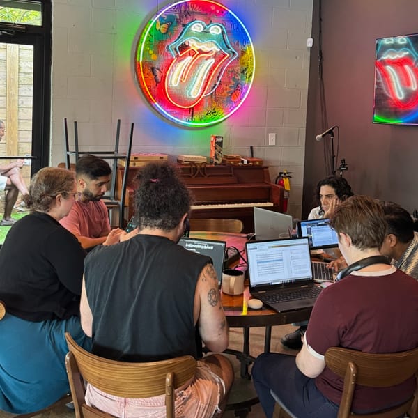 People working on laptops around a table, with a neon Rolling Stones sign on the wall.