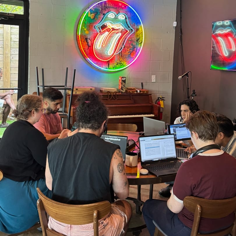 People working on laptops around a table, with a neon Rolling Stones sign on the wall.