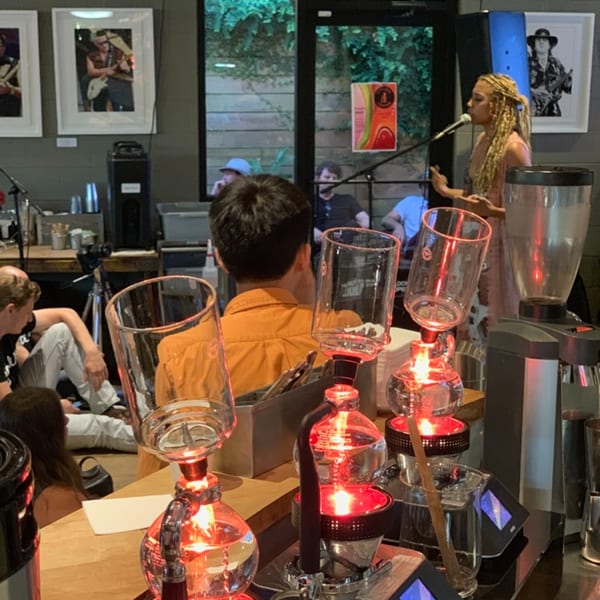 A woman with blonde dreadlocks sings into a microphone, with coffee brewing equipment in the foreground.