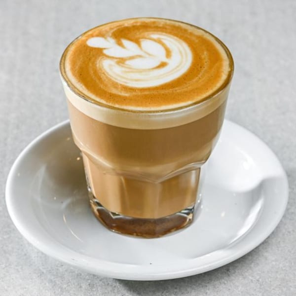 A glass of latte with a leaf design in the foam, on a white saucer.