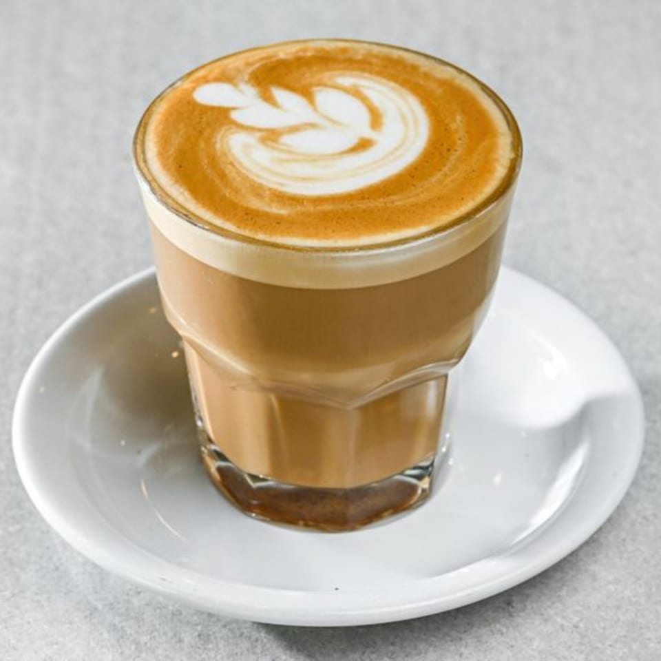 A glass of latte with a leaf design in the foam, on a white saucer.