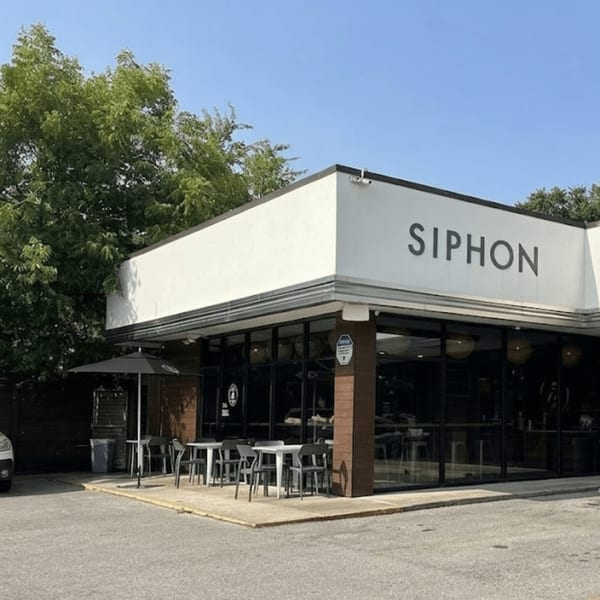Exterior of Siphon restaurant with outdoor seating under an umbrella, surrounded by trees and a clear sky.