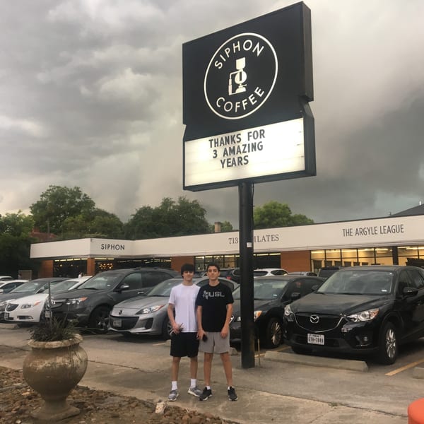 Two young men stand in front of a Siphon Coffee sign that reads "Thanks for 3 Amazing Years".