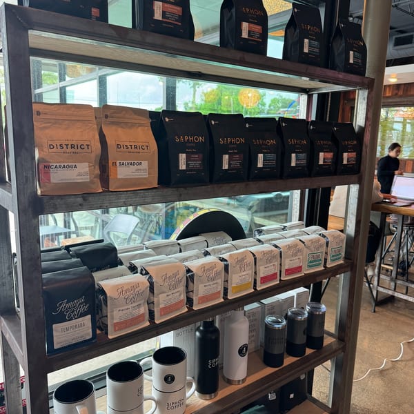 A metal shelf unit displays various brands of coffee bags, mugs, and tumblers in a cafe.