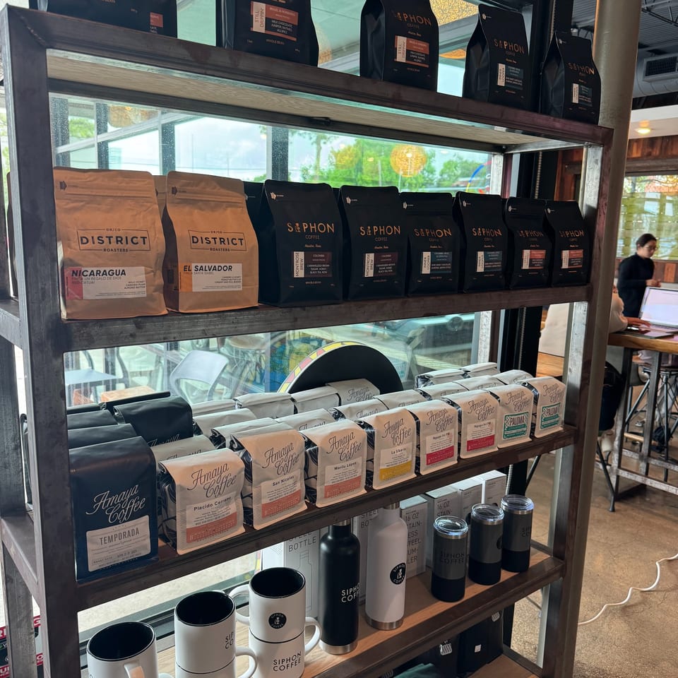 A metal shelf unit displays various brands of coffee bags, mugs, and tumblers in a cafe.