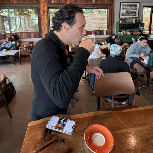 Man sipping coffee at a cafe table with other patrons and a phone displaying a coffee image.