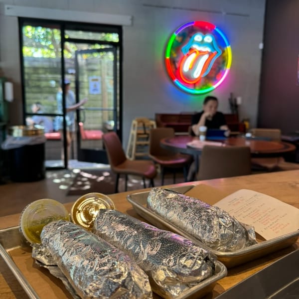 Foil-wrapped burritos and sauces on a tray, with a neon Rolling Stones sign and diner in the background.
