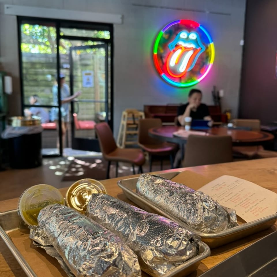 Foil-wrapped burritos and sauces on a tray, with a neon Rolling Stones sign and diner in the background.