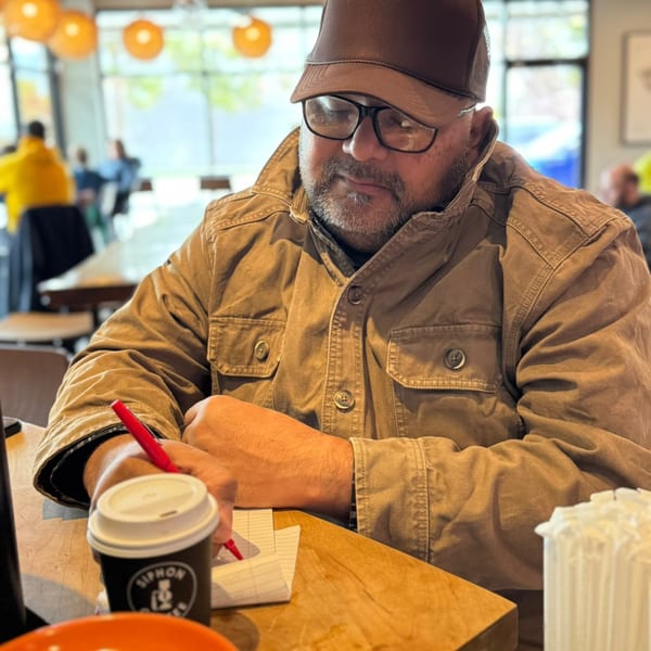 Man in a brown jacket and cap writing in a notebook with a coffee cup nearby.