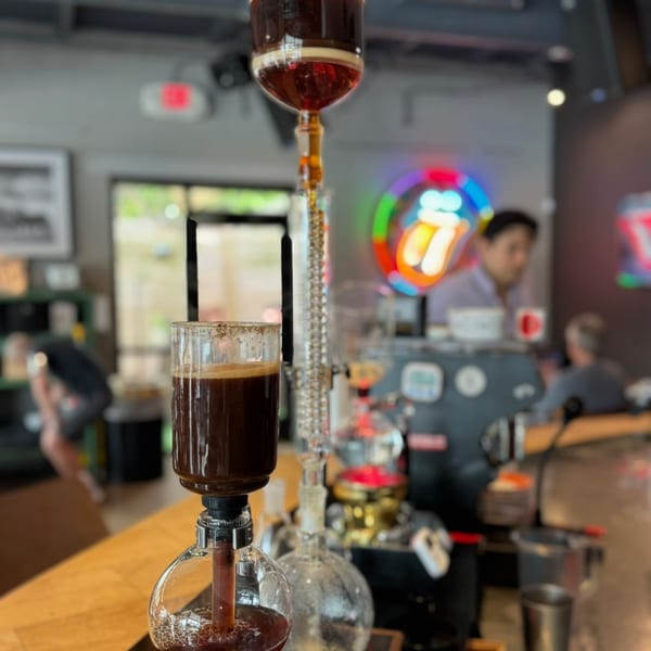 Coffee brewing in a siphon coffee maker on a wooden counter with blurred people and neon signs.