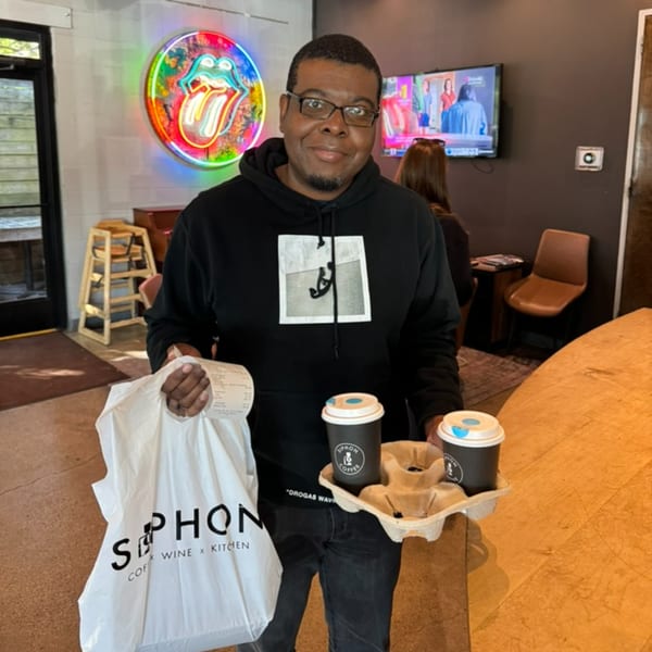 Man holding a Siphon Coffee bag and two coffee cups in a cafe with a neon Rolling Stones sign.