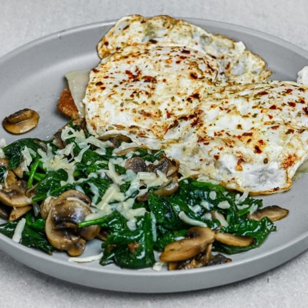 A gray plate holds two fried eggs with seasoning, sautéed spinach, mushrooms, and shredded cheese.