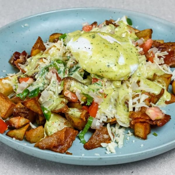 A blue plate filled with crispy potatoes, bacon, cheese, tomatoes, and a poached egg with green sauce.