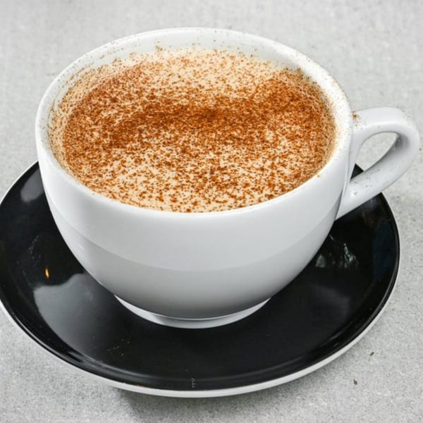 A white mug of frothy coffee, generously dusted with cinnamon, sits on a black saucer.